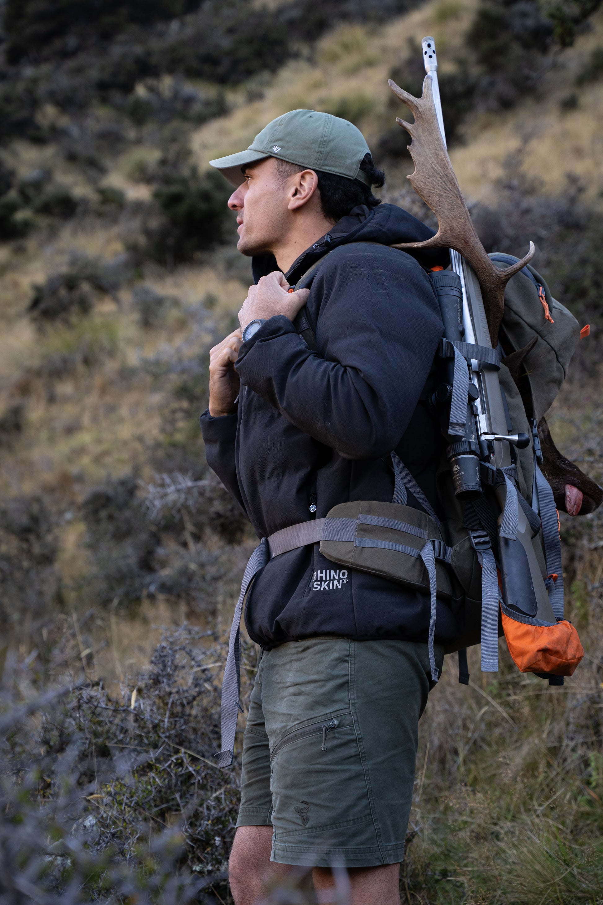 Man in outdoor setting with backpack and rifle, surrounded by nature.