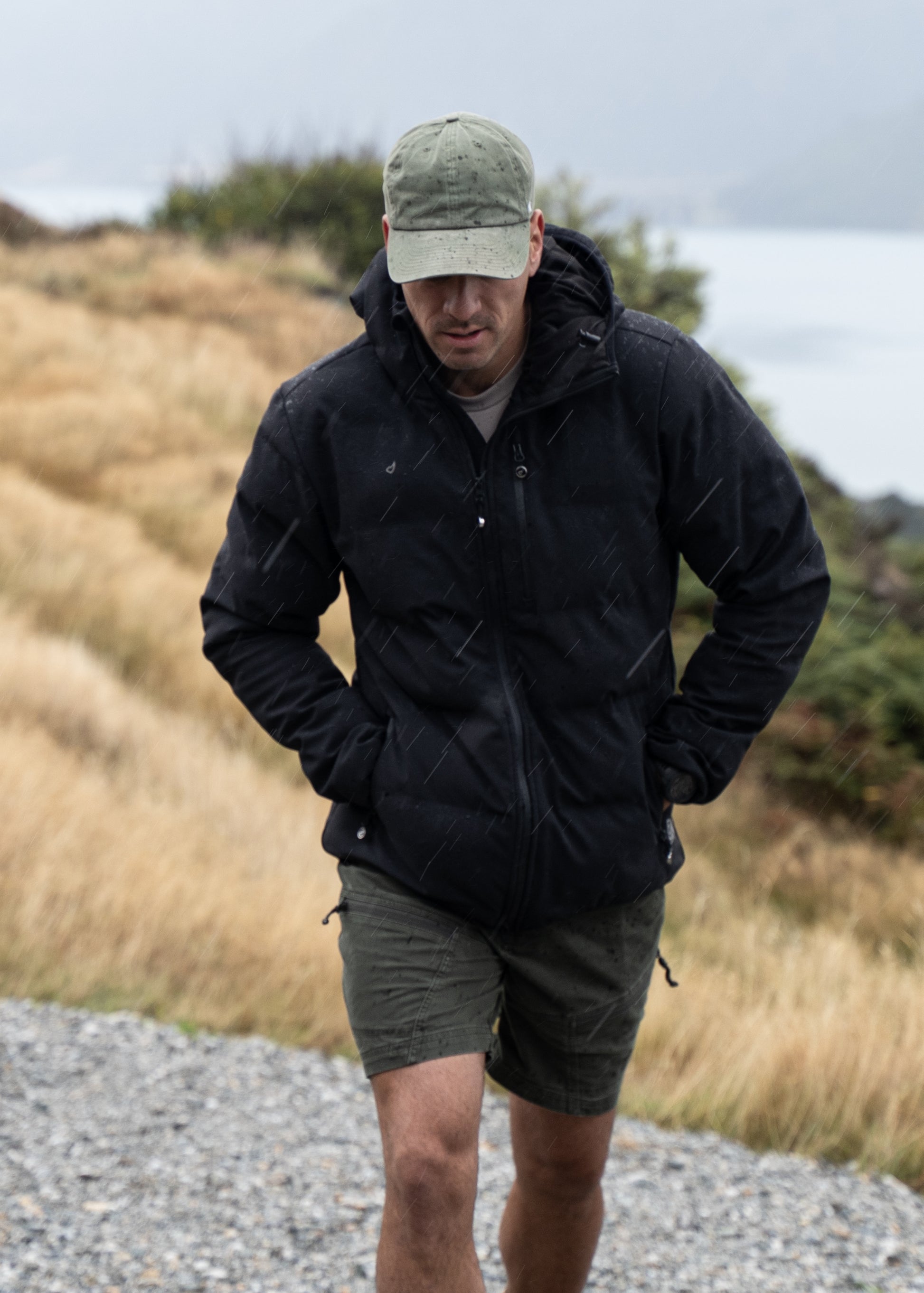 Man wearing a black jacket and cap standing on a gravel path with grass and water in the background