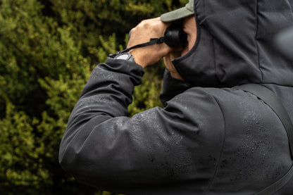 Person wearing a dark jacket with a hood, holding binoculars to their eyes, against a blurred green background.