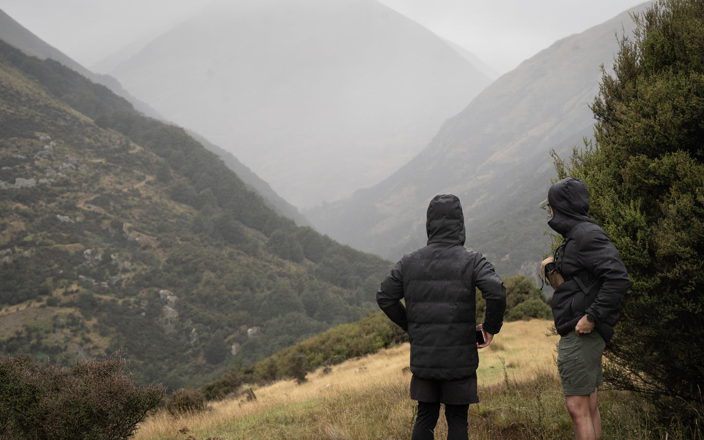Two people in dark jackets standing on a grassy hill with mountains in the background