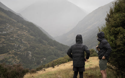 Two people in dark jackets standing on a grassy hill with mountains in the background