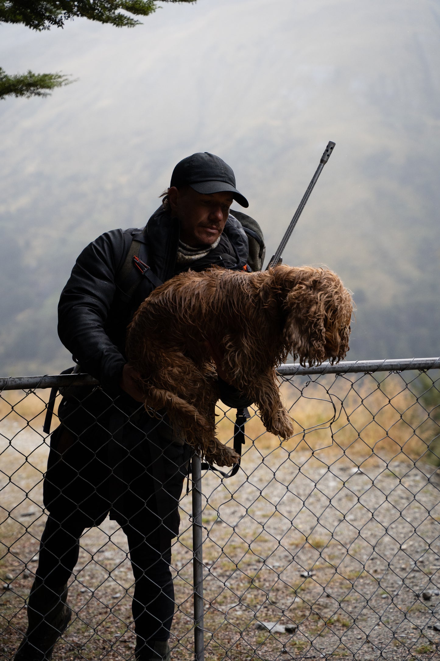 Man holding a dead animal behind a chain-link fence with a misty landscape in the background
