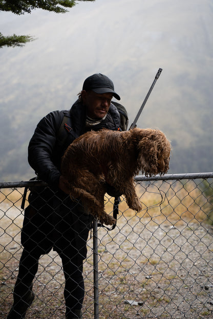 Man holding a dead animal behind a chain-link fence with a misty landscape in the background